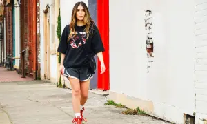 Donna con maglia over nera e pantaloncini sportivi cammina sul marciapiede, con edifici in mattoni e un muro bianco sullo sfondo.