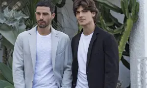 Due uomini eleganti indossano giacche con collo sciallato, una grigia chiara e una nera, con t-shirt bianche sotto, su uno sfondo verde.