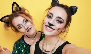 Due amiche si divertono a sperimentare come truccarsi a Carnevale con un look da gatta, baffi e orecchie nere scintillanti su sfondo giallo.