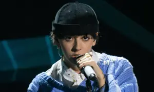 Un ariete cantante con un berretto nero e un maglione blu canta al microfono, con un'espressione intensa.