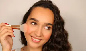 Una donna sorride mentre applica il fard trucco sulle guance con un pennello.