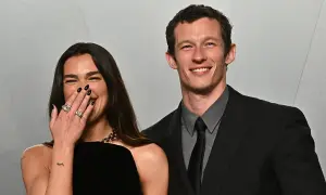 Dua Lipa ride con il suo compagno, Callum Turner, entrambi sorridenti. Lei copre la bocca con la mano, mostrando anelli scintillanti.