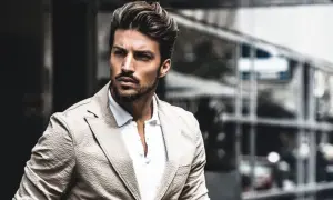 Mariano Di Vaio, con un blazer beige e camicia bianca, guarda intensamente verso l'esterno, con un'espressione pensierosa.