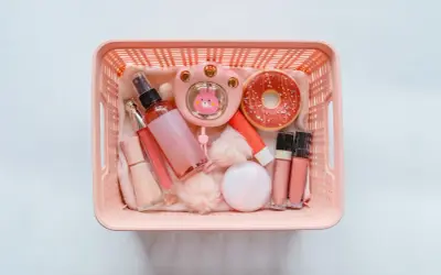 Cesto rosa pieno di trucchi: spray, lucidalabbra, fondotinta, ventilatore a forma di coniglietto, ciambella e altri accessori. Perfetti per viaggiare, i trucchi si possono portare in aereo!