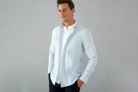 Uomo con camicia a righe azzurre e bianche, con colletto alla coreana e una camicia bianca sotto. La camicia con tasche è aperta, mostrando un look casual.