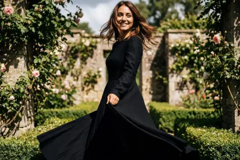 Donna sorridente in un abito con gonnellone lungo nero, che volteggia in un giardino fiorito con rose rosa e un muro di pietra.