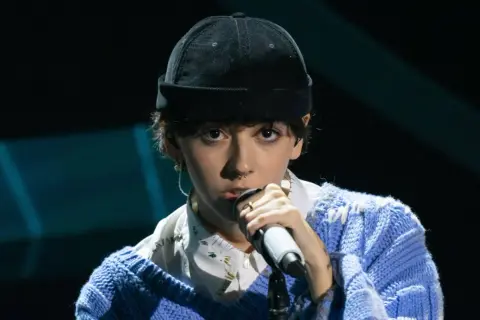 Un ariete cantante con un berretto nero e un maglione blu canta al microfono, con un'espressione intensa.