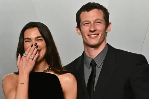 Dua Lipa ride con il suo compagno, Callum Turner, entrambi sorridenti. Lei copre la bocca con la mano, mostrando anelli scintillanti.