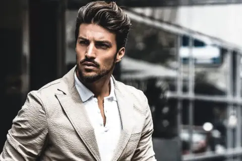 Mariano Di Vaio, con un blazer beige e camicia bianca, guarda intensamente verso l'esterno, con un'espressione pensierosa.