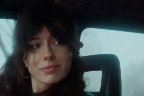 Ragazza con capelli scuri e orecchini a cerchio, guarda fuori dal finestrino, il cielo è celeste dalla porta.