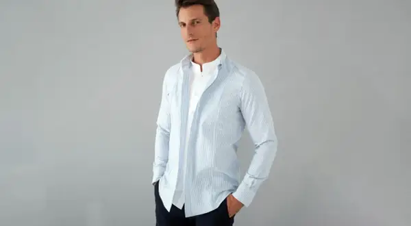 Uomo con camicia a righe azzurre e bianche, con colletto alla coreana e una camicia bianca sotto. La camicia con tasche è aperta, mostrando un look casual.