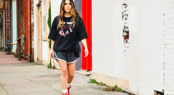 Donna con maglia over nera e pantaloncini sportivi cammina sul marciapiede, con edifici in mattoni e un muro bianco sullo sfondo.