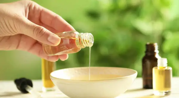 Mano versa un liquido dorato da una bottiglietta in una ciotola. È un olio per cuticole fatto in casa, perfetto per nutrire le unghie.
