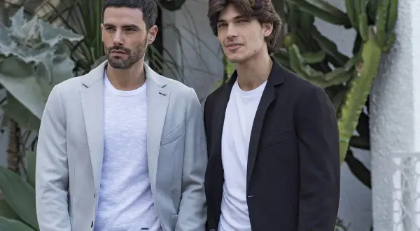 Due uomini eleganti indossano giacche con collo sciallato, una grigia chiara e una nera, con t-shirt bianche sotto, su uno sfondo verde.