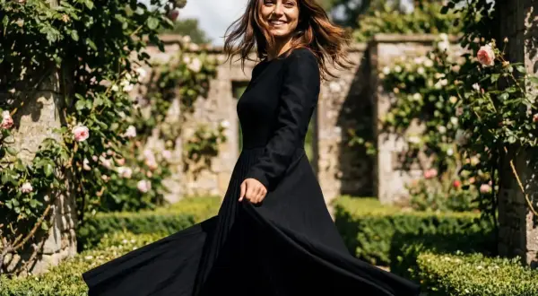 Donna sorridente in un abito con gonnellone lungo nero, che volteggia in un giardino fiorito con rose rosa e un muro di pietra.