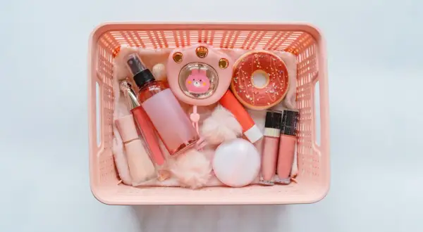 Cesto rosa pieno di trucchi: spray, lucidalabbra, fondotinta, ventilatore a forma di coniglietto, ciambella e altri accessori. Perfetti per viaggiare, i trucchi si possono portare in aereo!