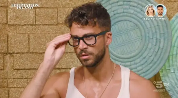 Rosario di Temptation Island, con occhiali neri, si sistema la montatura. Sullo sfondo, il logo del programma e le foto di Lucia e Rosario.