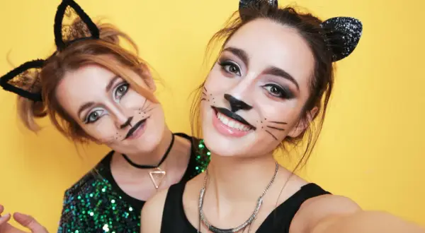 Due amiche si divertono a sperimentare come truccarsi a Carnevale con un look da gatta, baffi e orecchie nere scintillanti su sfondo giallo.