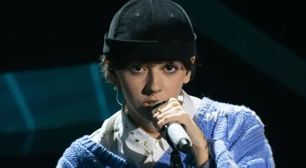 Un ariete cantante con un berretto nero e un maglione blu canta al microfono, con un'espressione intensa.