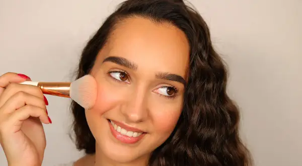 Una donna sorride mentre applica il fard trucco sulle guance con un pennello.