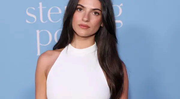 Ella Rubin, con lunghi capelli scuri e un abito bianco, posa con uno sfondo azzurro.