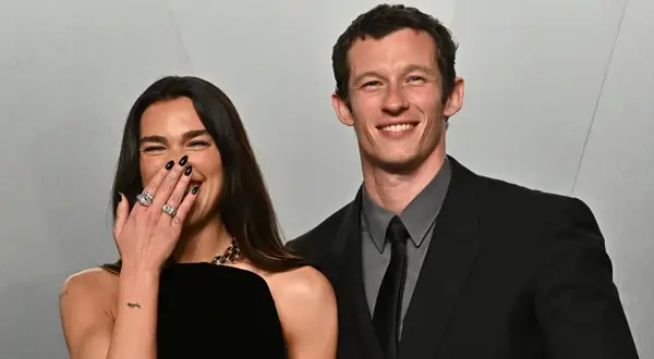 Dua Lipa ride con il suo compagno, Callum Turner, entrambi sorridenti. Lei copre la bocca con la mano, mostrando anelli scintillanti.
