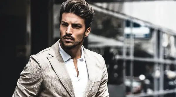 Mariano Di Vaio, con un blazer beige e camicia bianca, guarda intensamente verso l'esterno, con un'espressione pensierosa.