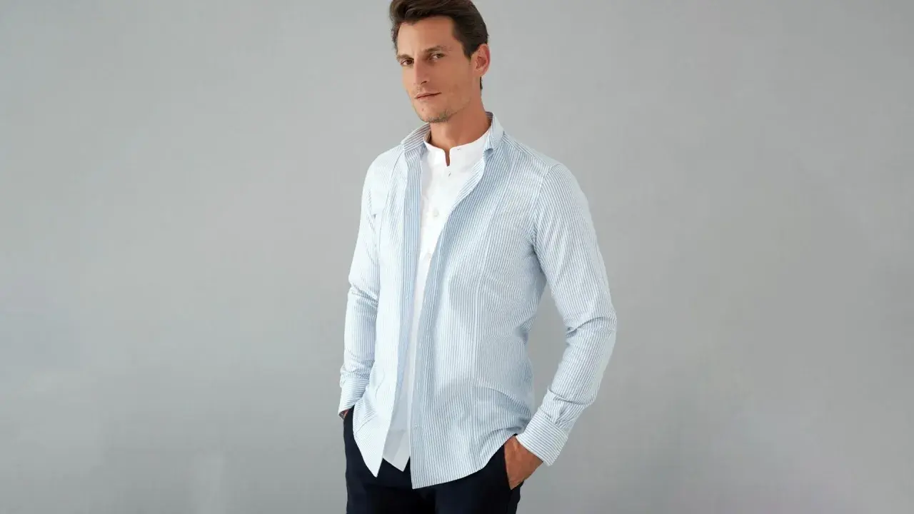 Uomo con camicia a righe azzurre e bianche, con colletto alla coreana e una camicia bianca sotto. La camicia con tasche è aperta, mostrando un look casual.