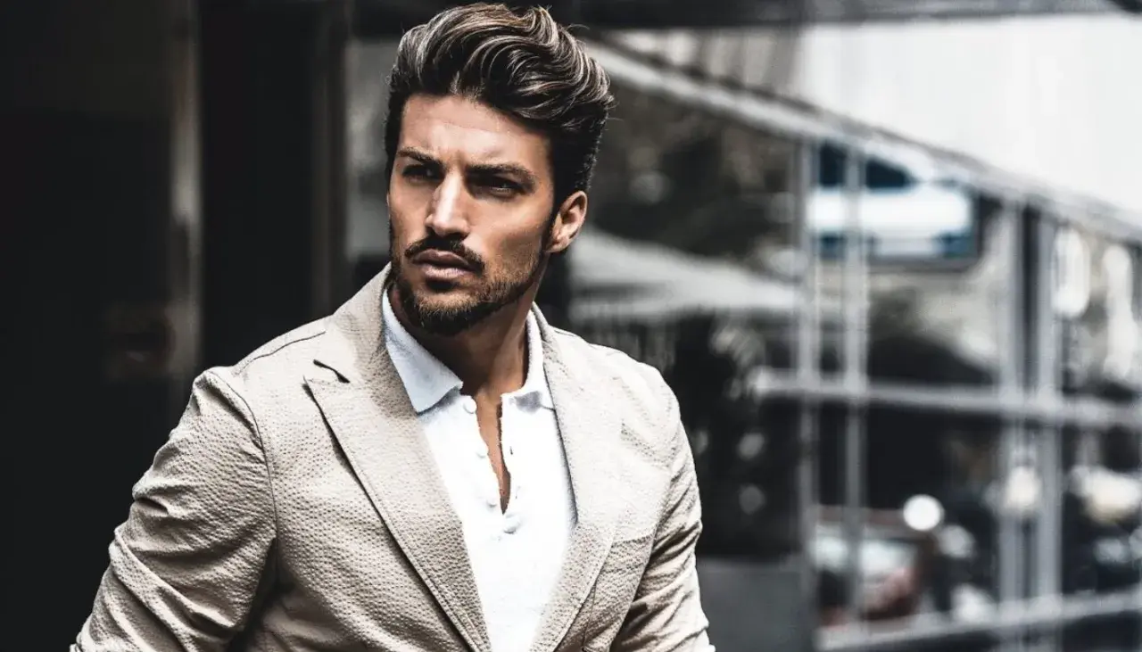 Mariano Di Vaio, con un blazer beige e camicia bianca, guarda intensamente verso l'esterno, con un'espressione pensierosa.