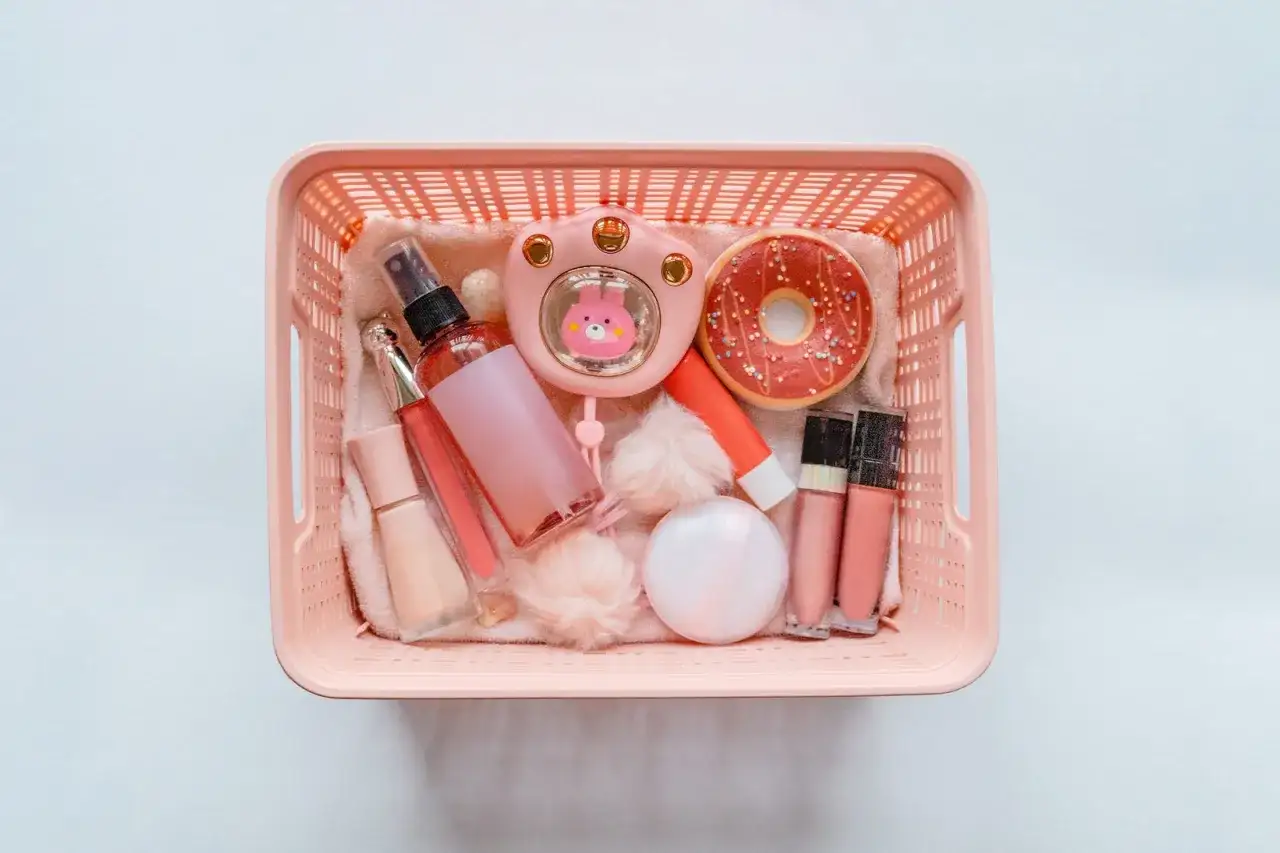 Cesto rosa pieno di trucchi: spray, lucidalabbra, fondotinta, ventilatore a forma di coniglietto, ciambella e altri accessori. Perfetti per viaggiare, i trucchi si possono portare in aereo!