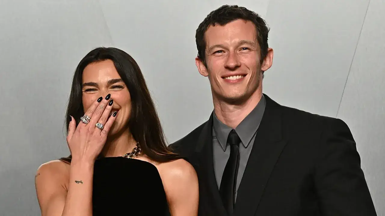 Dua Lipa ride e copre la bocca con la mano, mostrando anelli scintillanti. Accanto a lei, il suo compagno sorride.