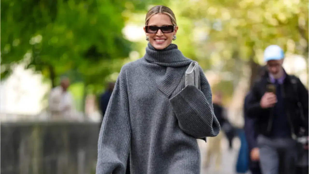 Donna sorridente con occhiali da sole e una comoda maglia over grigia a collo alto, con una borsa a tracolla.