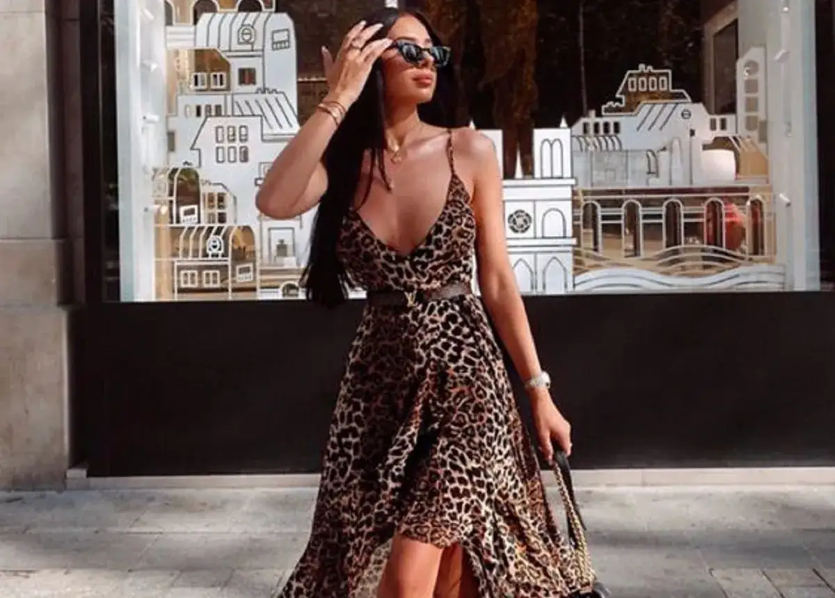 Donna con un elegante abito portafoglio leopardato, occhiali da sole e borsa, passeggia davanti a una vetrina con decorazioni architettoniche.