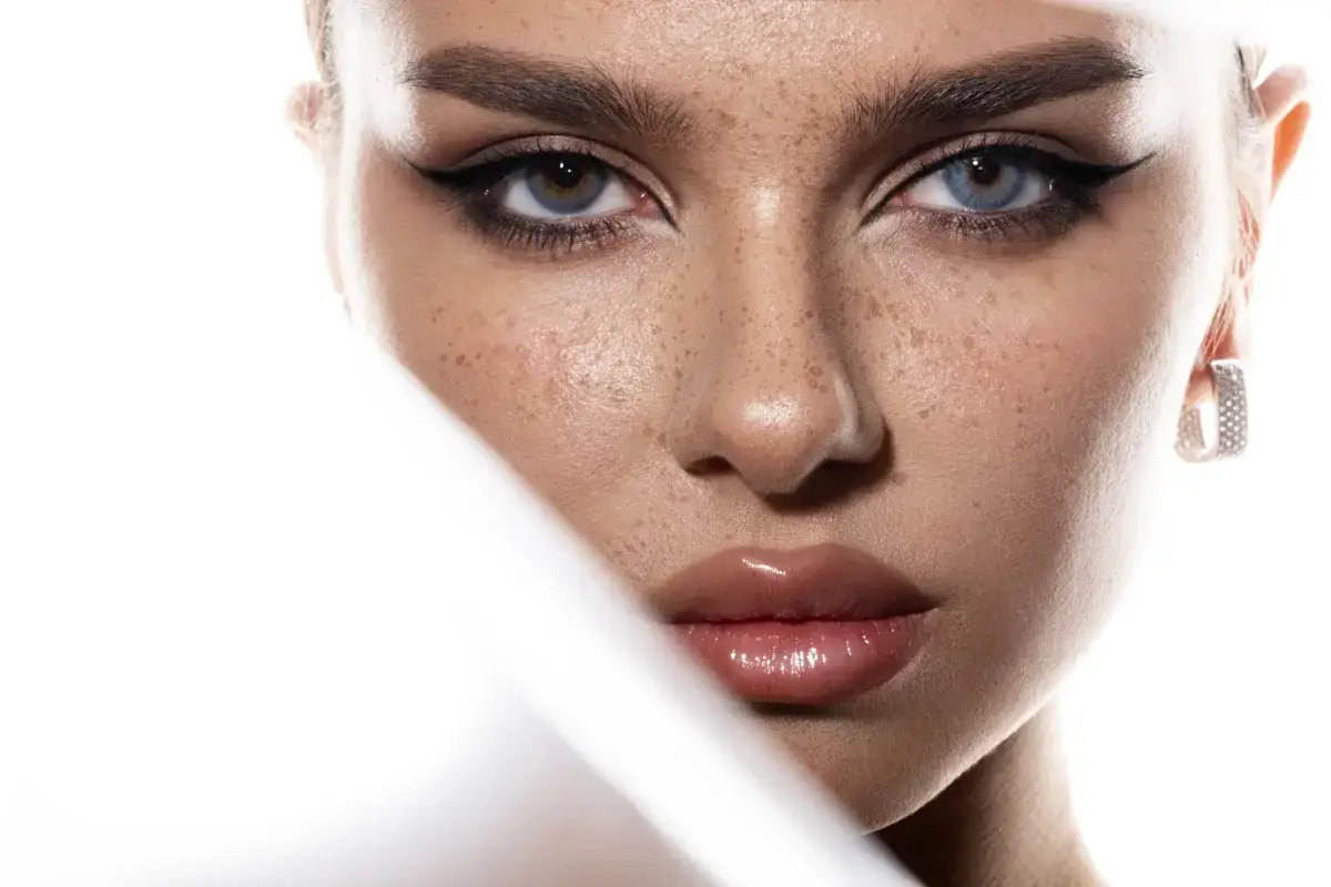 Close-up of a woman's face with freckles, dramatic winged eyeliner, and glossy lips.