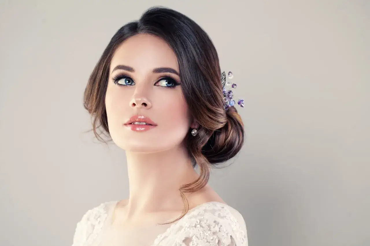 Woman with natural makeup, styled hair with purple hair accessory, and a white lace top.