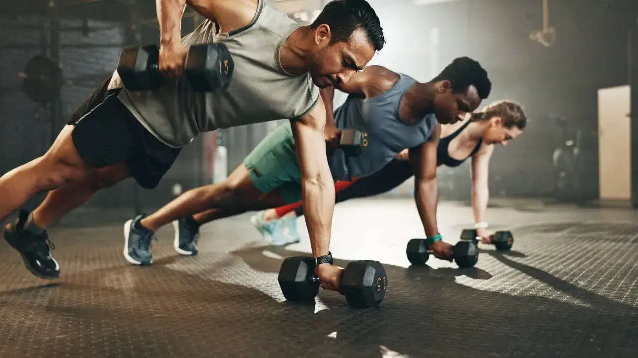 Uomini e donne in palestra eseguono un esercizio di plank con manubri, dimostrando i benefici dell'allenamento funzionale per forza e coordinazione.