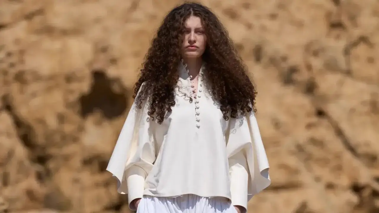 Modella con folti capelli ricci, indossa una blusa bianca con dettagli in pizzo e pantaloni ampi. L'ispirazione &egrave; a una moderna Athena Accorsi oggi.
