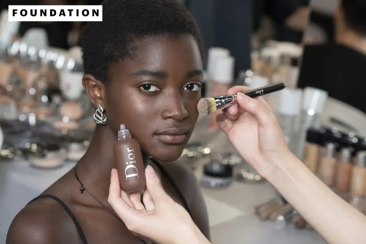 Modella con pelle scura riceve applicazione di fondotinta. Il correttore deve essere pi&ugrave; chiaro del fondotinta per un look impeccabile.