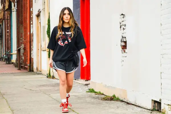 Donna con maglia over nera e pantaloncini sportivi cammina sul marciapiede, con edifici in mattoni e un muro bianco sullo sfondo.