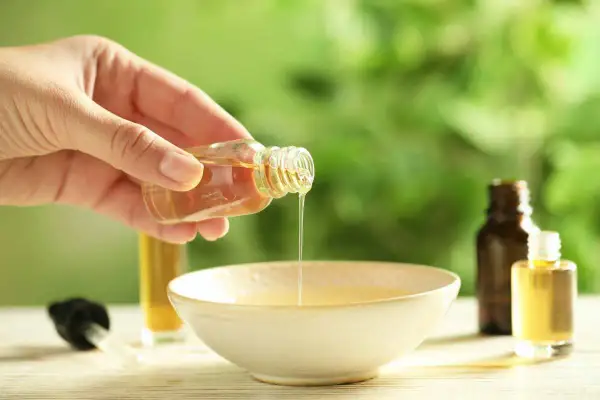 Mano versa un liquido dorato da una bottiglietta in una ciotola. È un olio per cuticole fatto in casa, perfetto per nutrire le unghie.