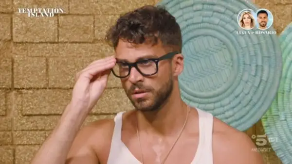 Rosario di Temptation Island, con occhiali neri, si sistema la montatura. Sullo sfondo, il logo del programma e le foto di Lucia e Rosario.