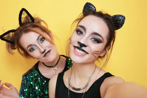Due amiche si divertono a sperimentare come truccarsi a Carnevale con un look da gatta, baffi e orecchie nere scintillanti su sfondo giallo.