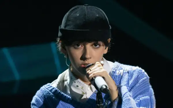 Un ariete cantante con un berretto nero e un maglione blu canta al microfono, con un'espressione intensa.