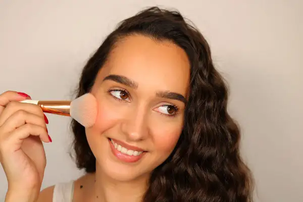 Una donna sorride mentre applica il fard trucco sulle guance con un pennello.